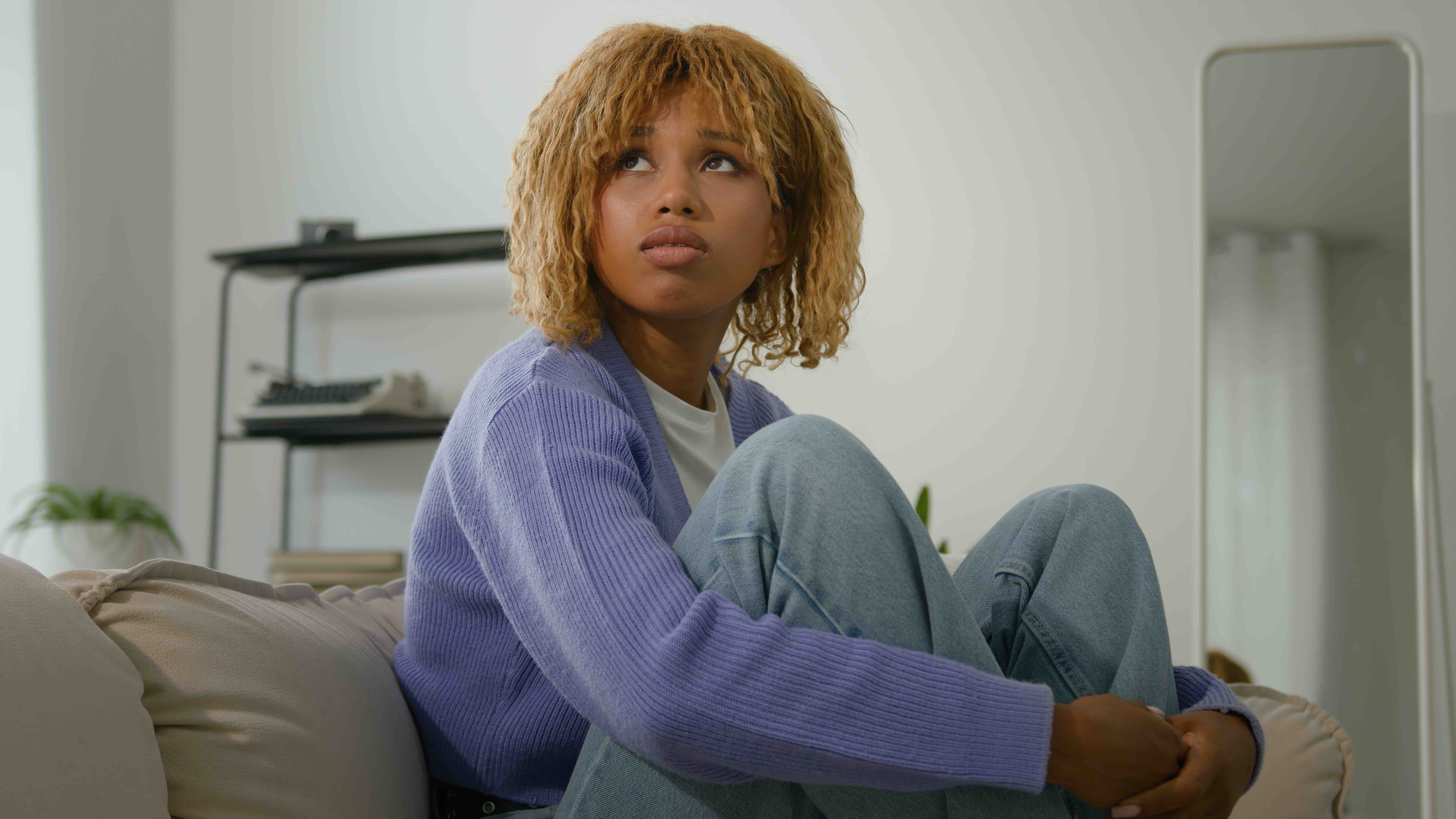 African American woman sad thoughtful young ethnic girl hug embrace knees lonely pensive female looking away thinking of problems sit alone at home upset stress depressed menstrual pain think suffer Antisociale persoonlijkheidsstoornis dame op de bank
